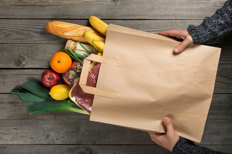 Man holding bag full of different healthy food on wooden kitchen table, top view