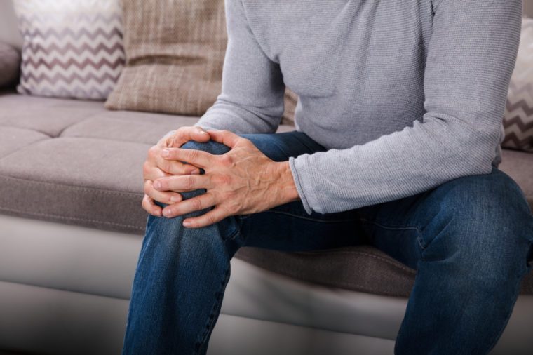 Man Suffering From Knee Pain Sitting On Sofa