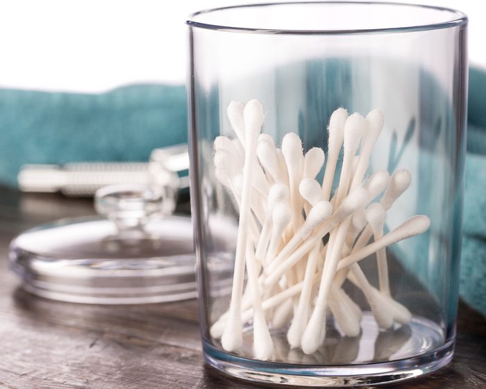 Cotton swabs in clear plastic jar in bathroom setting with a double edged razor and towel in the background on dark wood