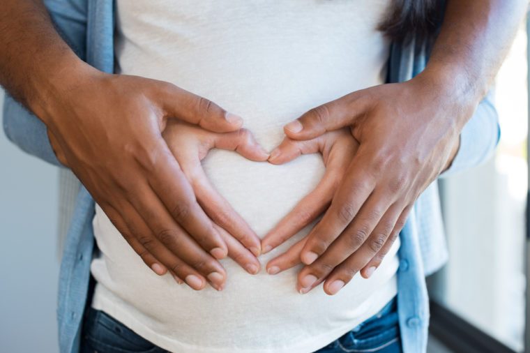 Black man holding belly of his pregnant wife making heart. Pregnant woman and loving husband hugging tummy at home. Heart of hands by multiethnic couple on pregnant belly.