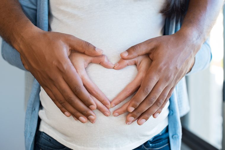 Black man holding belly of his pregnant wife making heart. Pregnant woman and loving husband hugging tummy at home. Heart of hands by multiethnic couple on pregnant belly.