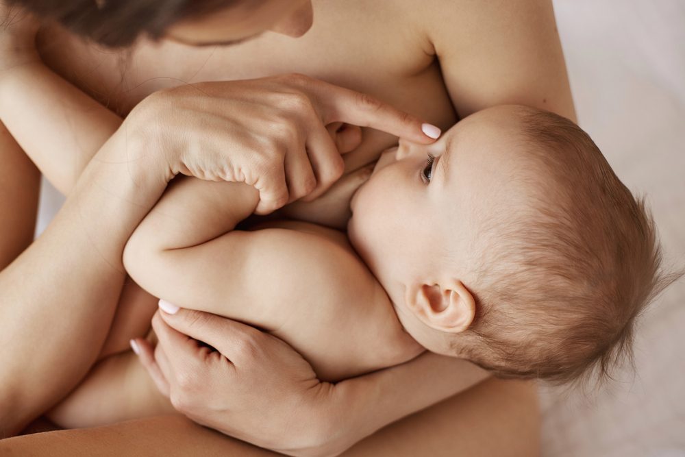 Young tender nude mother breastfeeding hugging her newborn baby sitting in bed at morning.