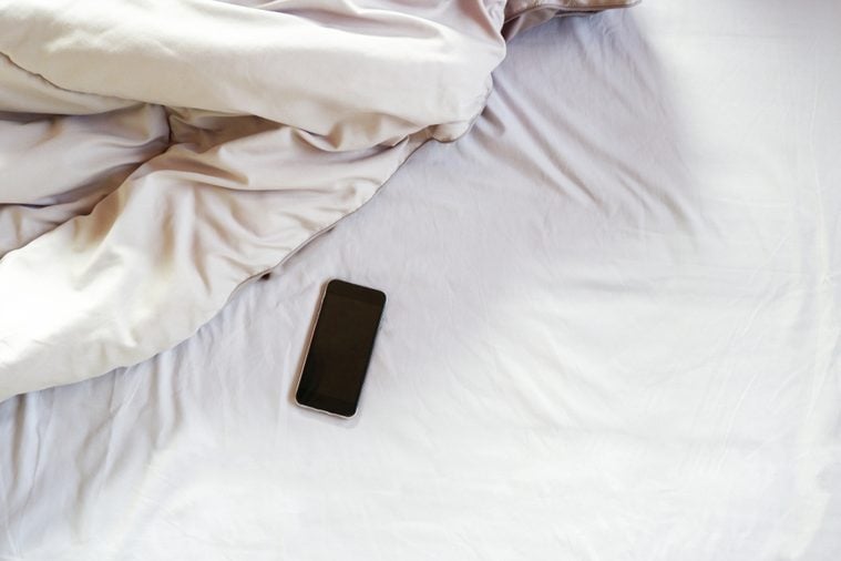 8 lugares donde nunca debes guardar o dejar tu teléfono| Vista superior de un teléfono inteligente negro en el pliegue de una sábana deshecha en el dormitorio