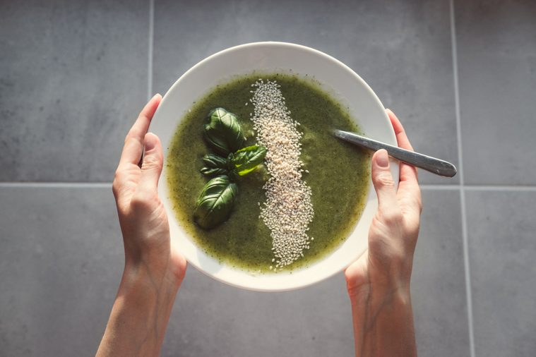 Fresh green healthy vegan soup in woman's hands on a grey tile background. Overhead shot of fresh soap in the hands. Broccoli soup with basil. 