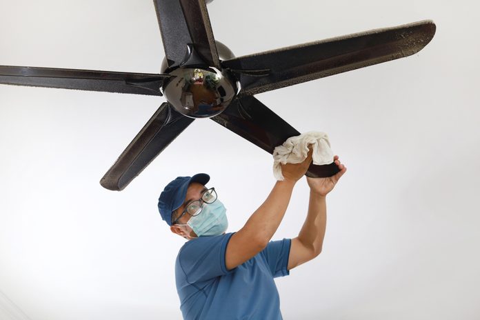 Asian Man Wearing A Protective Mask Cleaning Ceiling Fan At Home