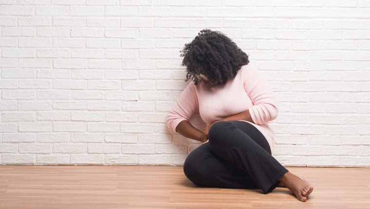Young african american woman sitting on the floor at home with hand on stomach because nausea, painful disease feeling unwell. Ache concept.