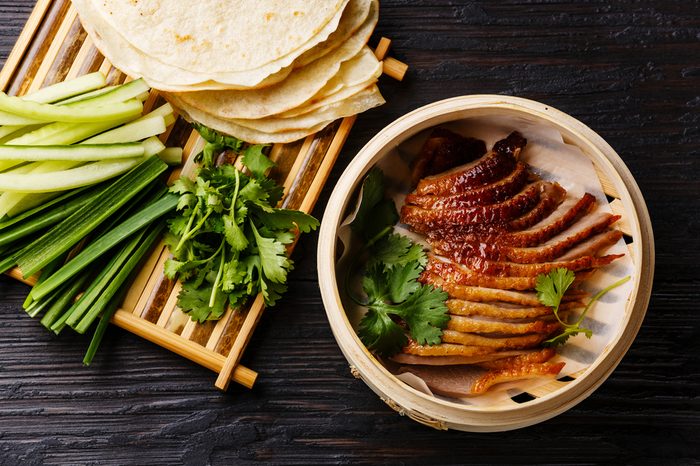 Peking Duck in bamboo steamer served with fresh cucumber, green onions, cilantro and roasted wheaten chinese pancakes on black burned wooden background