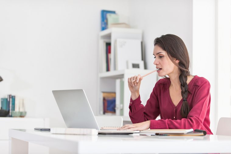 Portrait of a dark braided hair woman chewing her pencil while working home on her laptop. She is sitting at a big white table in a luminous place, wearing casual clothes. Focus on the woman
