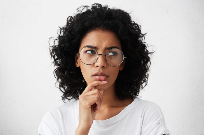 Doubtful young woman wearing trendy round eyeglasses frowning and looking sideways with indecisive and perplexed facial expression, touching her chin while can't decide what to wear on party