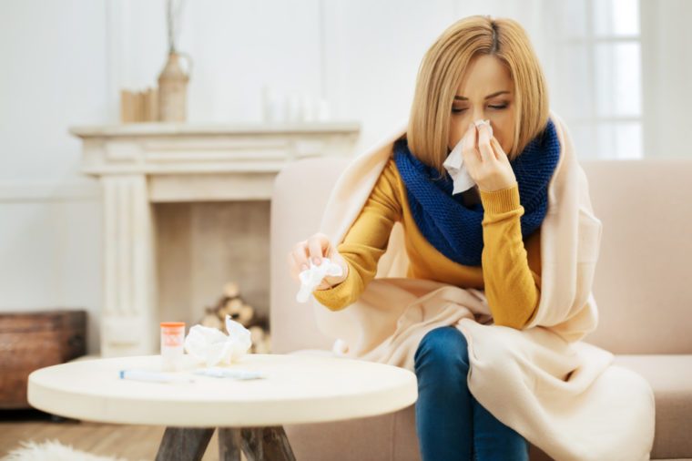Runny nose. Ill young blond woman having fever and blowing her nose while having a blanket on her shoulders and sitting on the couch with her eyes closed and table with pills in front of her