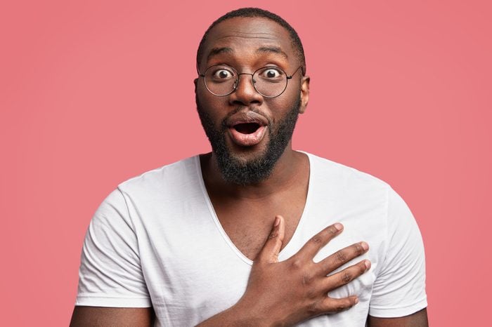 Studio shot of excited glad African American male model keeps hand on chest, looks with surprise and amazement, doesn`t expect to recieve congratulation from colleagues. Pleased dark skinned guy