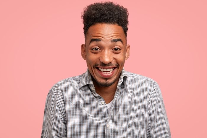 Cheerful dark skinned guy has trendy haircut, giggles happily, dressed in checkered shirt, stands against pink background, models in studio. Smiling African American man expresses positive emotions