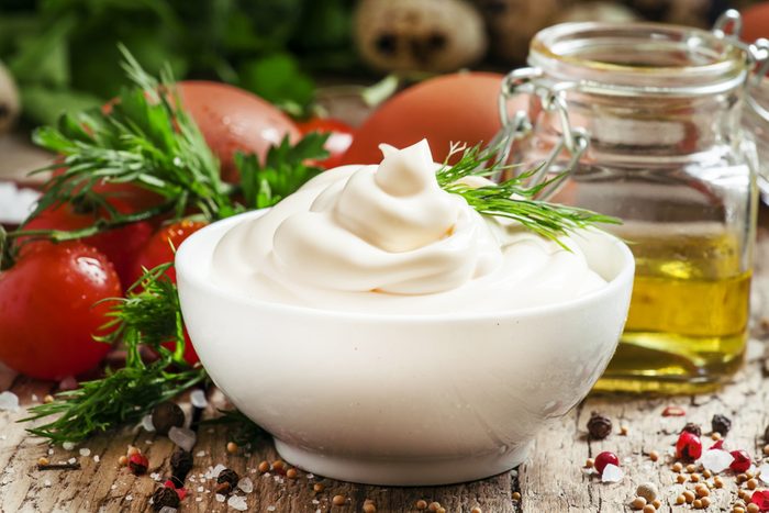 Homemade mayonnaise sauce in a white bowl, jar with olive oil, eggs, salt, spices, mustard, herbs, cherry tomatoes on old wooden background, selective focus