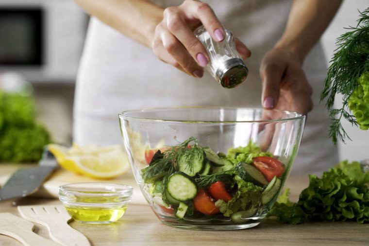 Woman adding salt in vegetable salad glass bowl, health care, excessive salting