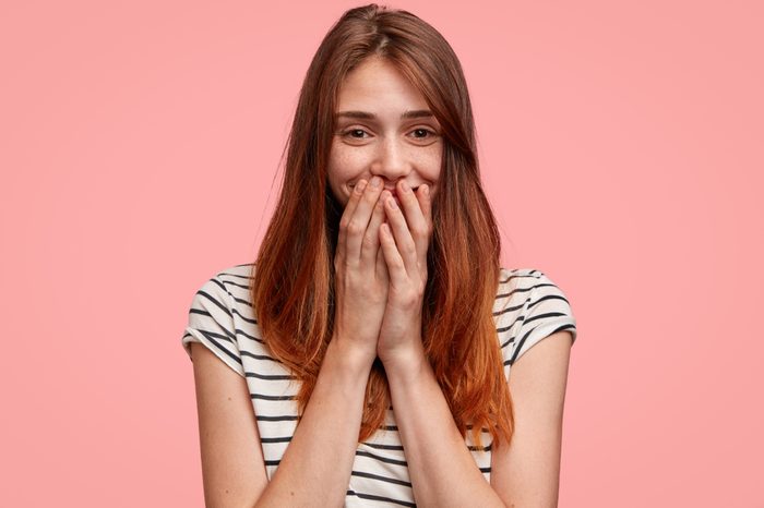 Satisfied European female laughs happily, covers mouth as tries to stop giggling, wears casual striped t shirt, poses against pink background. Beautiful young woman has long hair and freckles