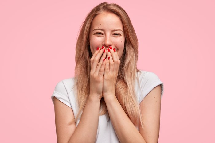 Horizontal shot of good looking young female giggles joyfully, covers mouth as tries stop laughing, wears casual white t shirt isolated on pink background. Happy woman recieves proposal from boyfriend