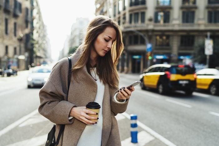 Travel blogger using route application on the mobile phone to find the needed address in a city. Young stylish blonde woman reading emails on the smartphone while passing by with a take away coffee