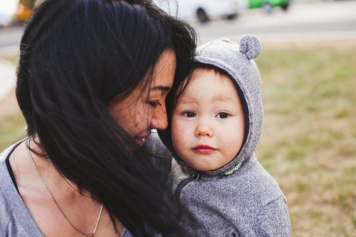 Beautiful young asian woman with freckles and her son outdoors. Mother brunette with dark hair holds her blond son in bear hood. Emotions on baby face. Unusual appearance, diversity, heredity concept