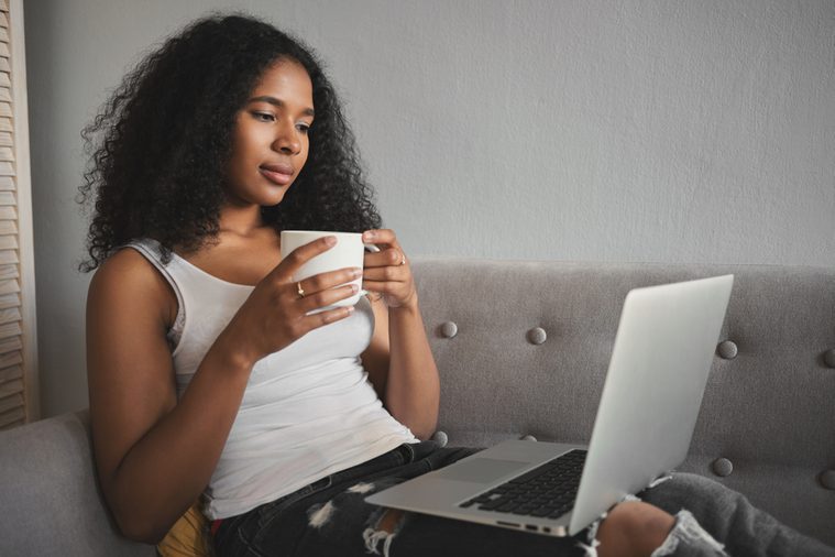 Sideways shot of fashionable attractive young dark skinned female in ripped jeans relaxing on couch with portable computer on her lap, drinking coffee and watching favorite tv series online