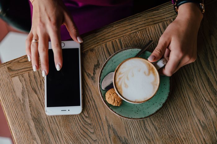 Female hands hold a cell phone and a cappuccino cup on a saucer with a cookie. Against the background of the table. white manicure and telephone. top view Black screen.