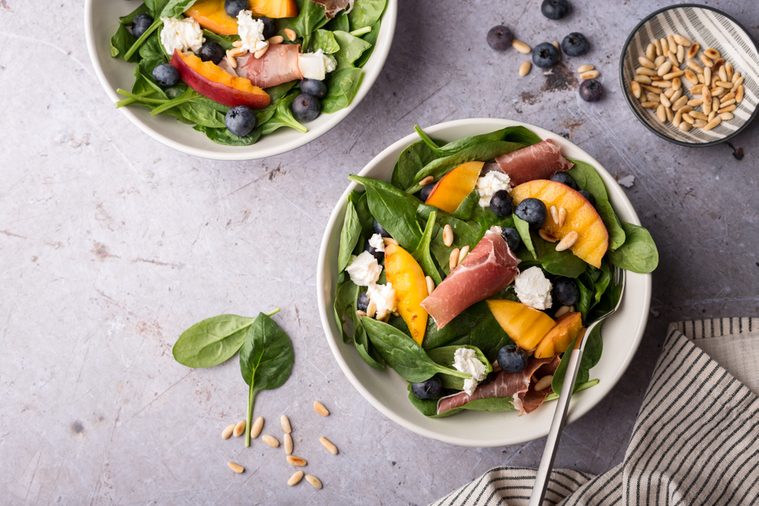 Grilled peach salad with prosciutto, blueberry, baby spinach, goat cheese and pine nuts on concrete background, overhead view, copy space