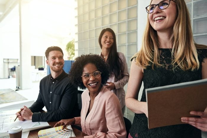 Laughing young businesswoman standing in a modern office with a diverse group of work colleagues smiling in the background