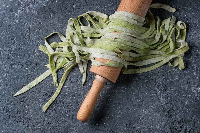 Fresh raw uncooked homemade green spinach pasta tagliatelle on wooden rolling pin over dark texture concrete background. Top view with space.