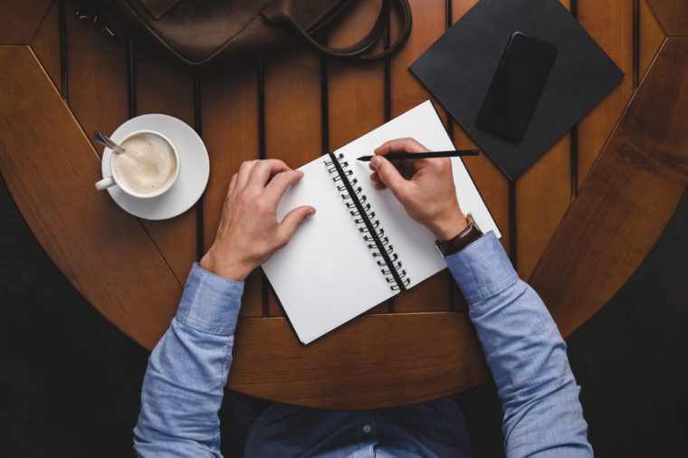 overhead view of man writing in notepad at wooden table with coffee and leather bag