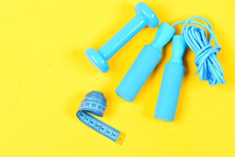 Gym and healthy lifestyle tools. Weight loss and sports concept. Dumbbell, twisted measuting tape and jumping rope lay top on yellow background. Centimeter in blue color near colorful sport equipment.
