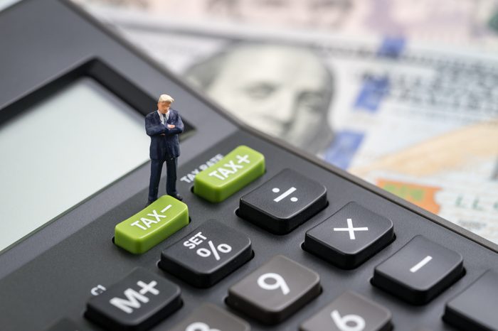 Tax cuts or reduce concept, miniature people business man president standing with TAX minus button on calculator with background of blurred US Dollar banknotes, United States government tax overhaul.