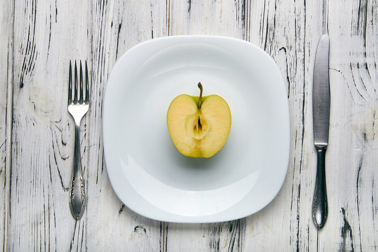 Eating for a girl, women on a strict diet. Half a green sweet and sour apple on a white plain plate. Too little food for losing weight. Anti-obesity diet by evil nutritionist.