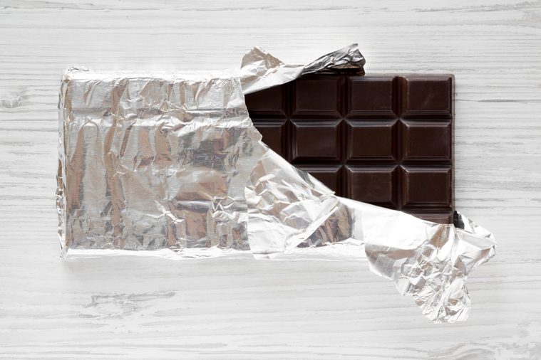 Dark chocolate in a wrapper over white wooden background, overhead view. Flat lay, from above, top view.