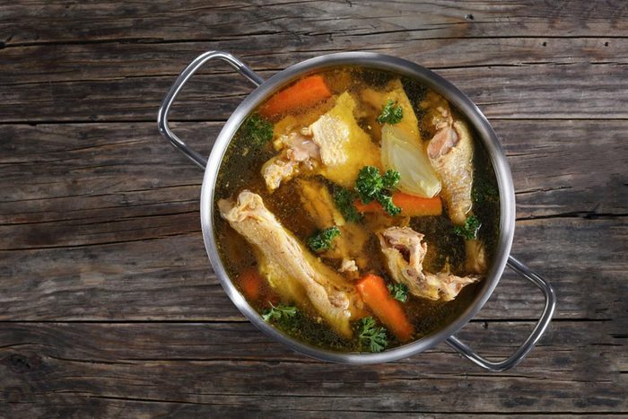 clear Chicken broth with pieces of rooster meat on bone and vegetables in a metal casserole on dark wooden table, view from above