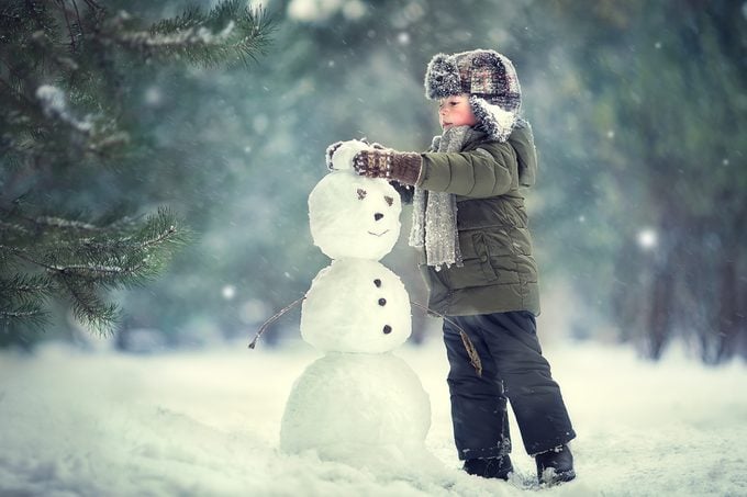 Cute little boy in earflaps hat is making a snowman. Image with selective focus and toning