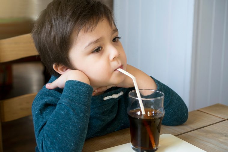 Cute toddler drinking cold drink, Little boy drink soda or soft drink through straw sitting on the chair in English pub, Child waiting for food with bored face