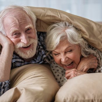 Cheerful old married couple lying in bed under blanket. Woman is laughing and man is looking at camera with smile. Concept of happiness