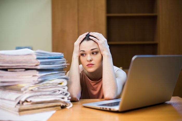 A young woman sits in front of a pile of papers and a computer holding her head