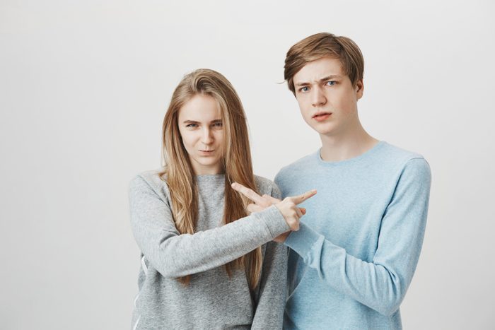 Siblings trying to accuse each other in front of angry mom. Serious unhappy couple with fair hair, frowning and pointing at each other, blaming and saying it is your fault, arguing over gray wall