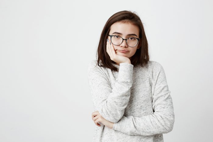 Shot of bored annoyed beautiful teenage girl with straight dark hair looking at camera with dark eyes holding hand under chin, wearing trendy eyewear and long-sleeved sweater, got tired at school