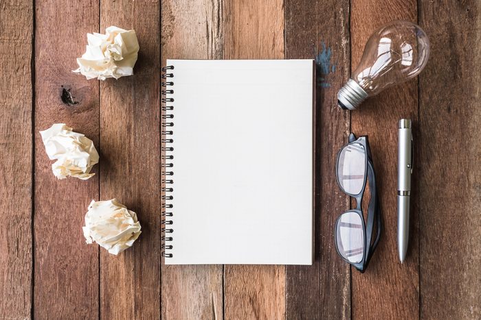Top view of notebook with crumpled paper balls, pen, glasses and light bulb on wooden table background.