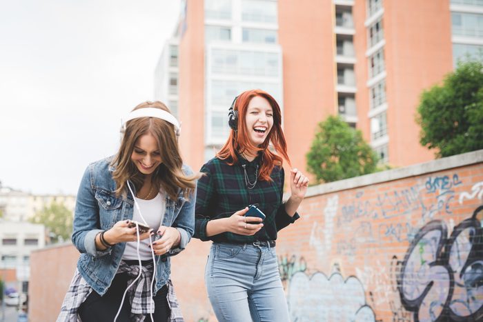 Knee figure of two young handsome caucasian blonde and redhead straight hair women dancing in the city listening music with headphones and smartphone, laughing - music, fun, freedom concept
