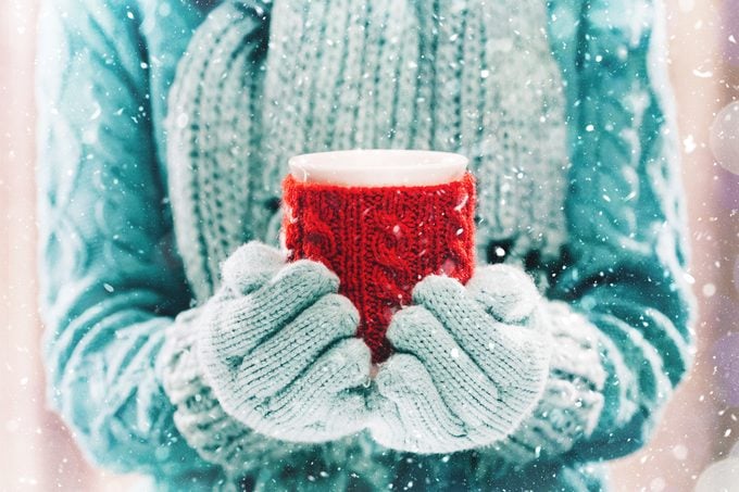 Woman holding winter cup close up on light background. Woman hands in teal gloves holding a cozy mug with hot cocoa, tea or coffee and a candy cane. Winter and Christmas time concept.