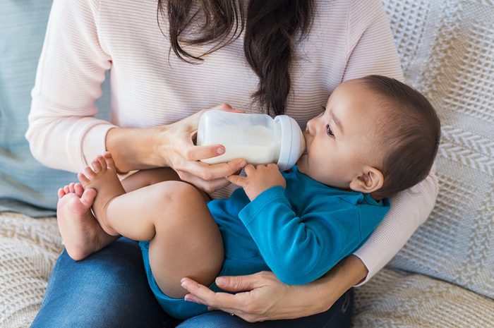 Little infant baby lying on mothers hand drinking milk from bottle. Hispanic loving mother feed her cute toddler while sitting on sofa.