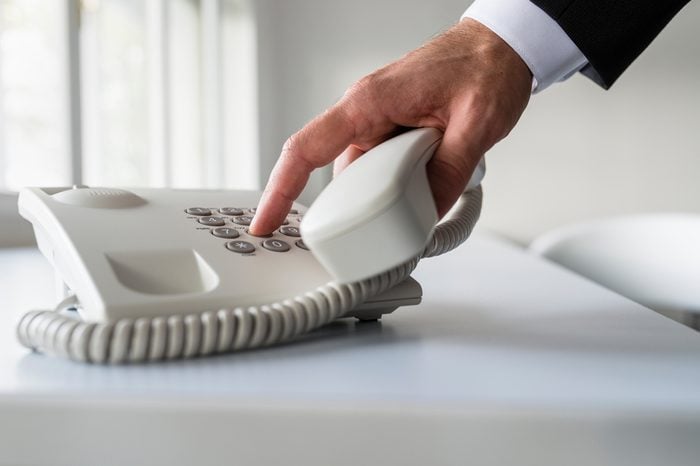 Male hand dialing a telephone number in order to make a phone call on a classical white landline telephone on an office desk.