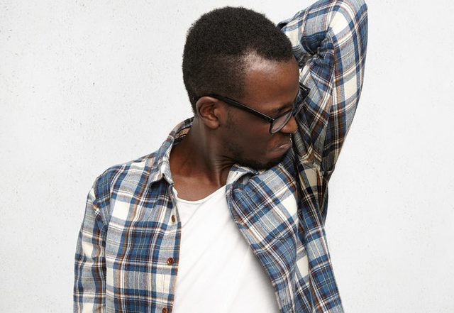 Disgusted young male wearing checkered shirt and glasses smelling wet sweaty armpit after stressful meeting, feeling nauseous, screwing lips. Black man can't stand bad smell. Hyperhidrosis and hygiene