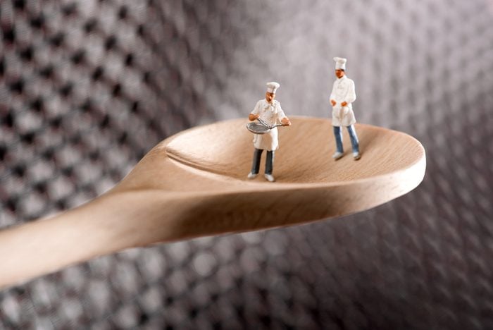 Two miniature figures of chefs or cooks on a wooden spoon over a textured metal background in a concept of catering and food preparation