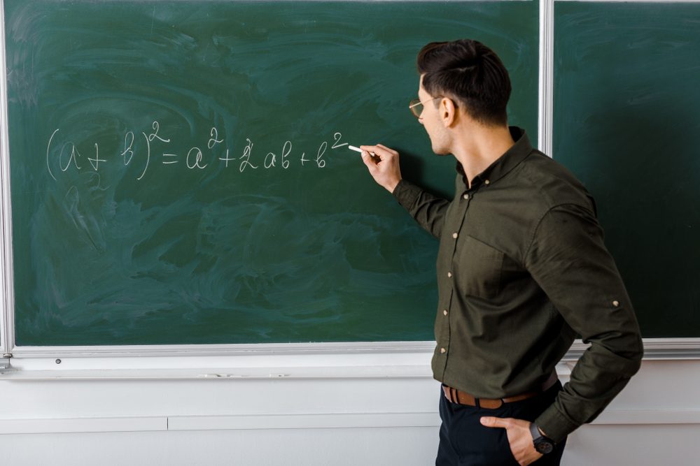 male teacher in formal wear writing equation on chalkboard in maths class