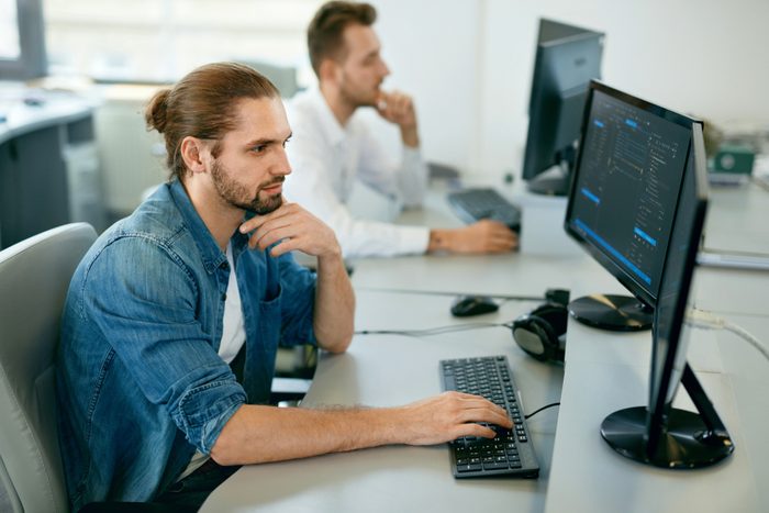 Programmers Working, Looking At Computer In IT Office. Handsome Young Men In Casual Closes Typing Codes, Working On Computer While Sitting At Workplace. High Quality Image.