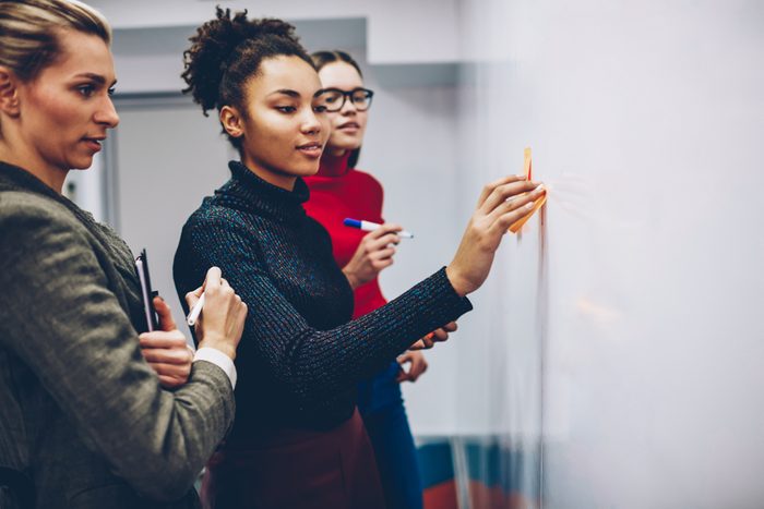 African american female student using stickers on flip chart for explaining ideas during lesson with business coach, multiracial crew of women employees having brainstorming session near board