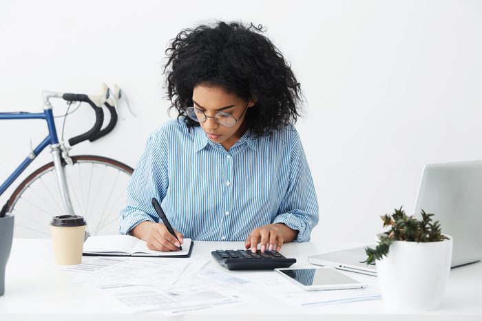 Portrait of attractive hardworking businesswoman with Afro hairstyle busy doing paperwork at office desk, working through finances, using calculator and making notes in her notebook with pen